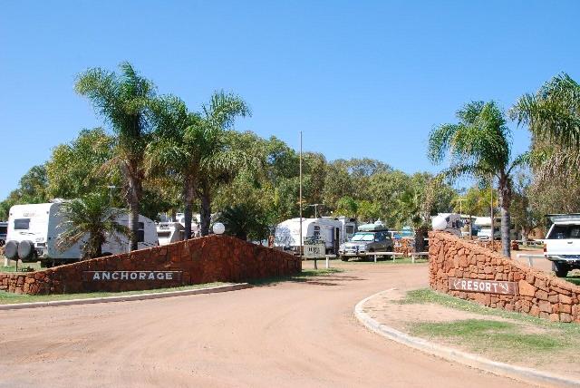 Entrance to Anchorage Caravan Park in Kalbarri, showing palm trees, caravans, and a wide gravel drive under blue skies.