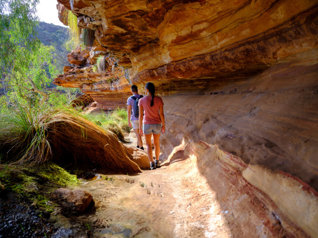 Two hikers walking along the riverbed section of the Kalbarri Loop Trail in winter, surrounded by ancient red sandstone gorge walls.